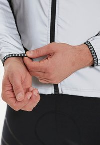 Lys grå cykeljakke med sort lynlås og manchetter. Manchetterne har et reflekterende accent. Hænder justerer ærmelængden, mens der bæres sorte shorts.