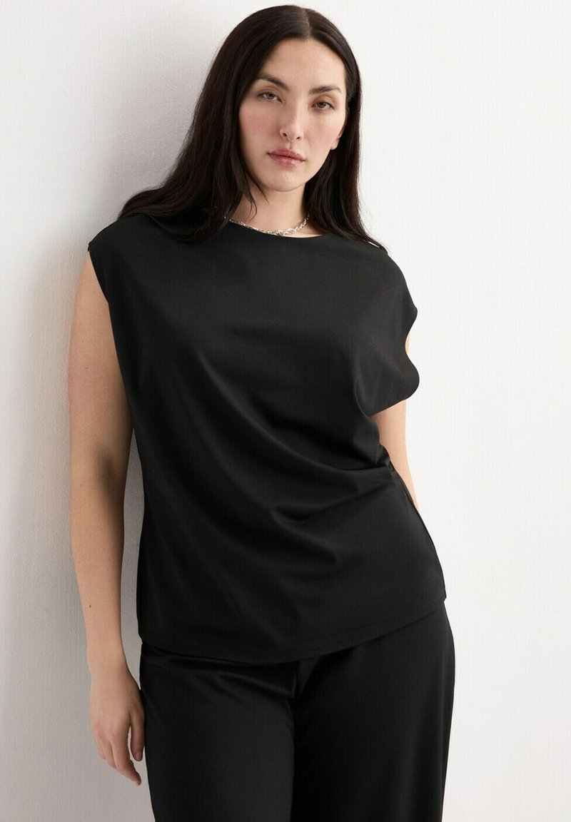 Femme aux longs cheveux bruns portant un haut noir sans manches et un pantalon noir, debout contre un mur blanc, regardant vers la caméra.