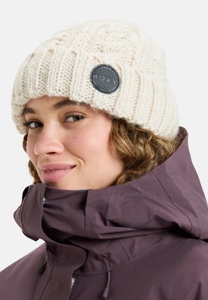 Gehaakte crèmekleurige beanie met een getextureerd patroon, voorzien van een zwart rond logo-embleem. Gecombineerd met een paars jack met hoge kraag.