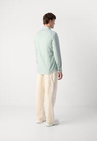 Cămașă lungă, verde deschis, cu guler cu nasturi, purtată cu pantaloni cargo bej, având buzunare laterale. Textură de țesătură netedă.