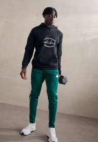 Felpa nera con logo bianco scritto, pantaloni della tuta verdi a gamba tapered con strisce laterali e sneakers bianche. Il modello tiene un manubrio.