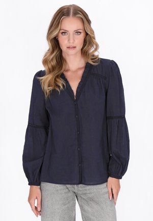 Femme aux cheveux blonds ondulés portant un chemisier bleu marine boutonné avec un col à volants et des manches longues bouffantes, assorti à un pantalon gris clair.