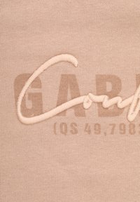 Tessuto beige con ricamo testurizzato con la scritta "GAB" e una linea, sovrapposto a una grafica stampata più chiara "IOS 49.798."