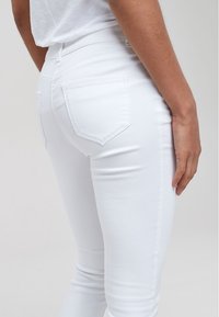 Persona che indossa jeans bianchi aderenti e una maglietta bianca, mostrata di spalle con un focus sui dettagli della tasca e della cucitura laterale dei jeans.