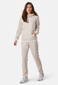 Completo felpa beige con pantaloni della tuta coordinati, dotato di tasca frontale e cappuccio con cordino. Tessuto morbido con branding minimo, abbinato a scarpe bianche.