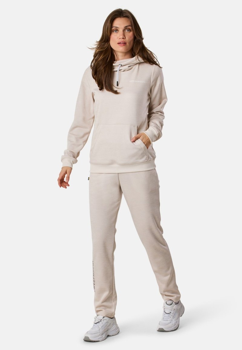 Kobieta stoi w beżowym dresie ze sweatshirta i dopasowanych spodni dresowych, w białych sneakersach, na jednolitym tle.