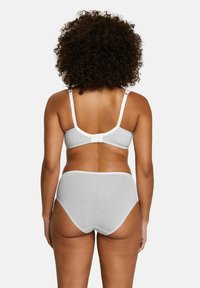 Femme aux cheveux bouclés vue de dos, portant un soutien-gorge gris clair et une culotte taille haute, sur fond blanc uni.