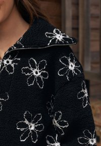 Sort fleece-pullover med høj krave, der har hvid blomsterbroderi og en sølvfarvet lynlås. Blød tekstur med et hyggeligt udseende.