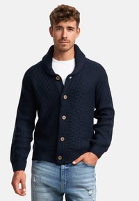 Cárdigan de punto azul marino con cuello chal, cuenta con botones de madera, tejido texturizado y puños acanalados. Combinado con jeans de lavado claro desgastados.