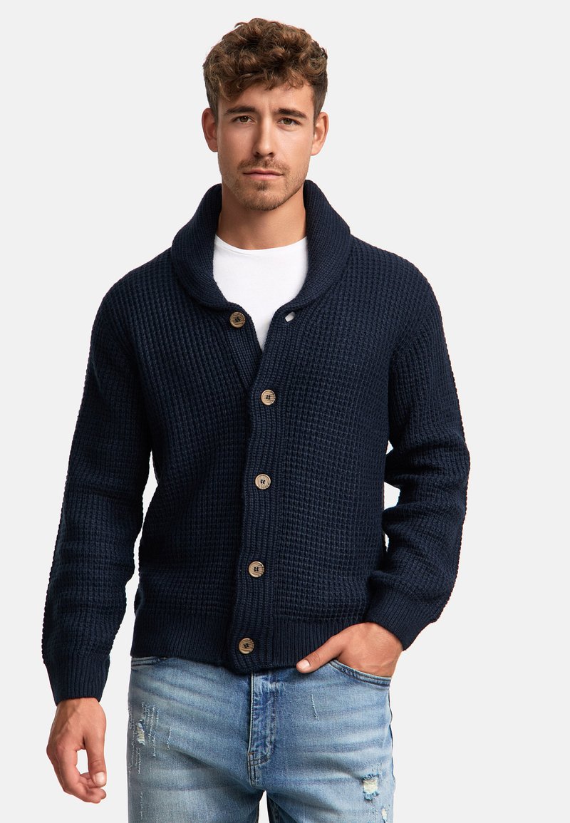 Cárdigan de punto azul marino con cuello chal, cuenta con botones de madera, tejido texturizado y puños acanalados. Combinado con jeans de lavado claro desgastados.