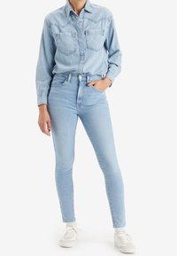 Camisa de mezclilla azul claro con dos bolsillos en el pecho, cierre de botones en la parte delantera y mangas largas. Combinada con jeans ajustados de tiro alto en azul claro.