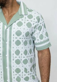 Chemise à manches courtes avec boutonnière en vert menthe et blanc, présentant des motifs géométriques, un col en V, et deux rayures blanches sur les manches.