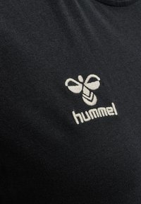 T-shirt nera realizzata in morbido tessuto, con un logo di ape ricamato in bianco e il testo "hummel" sottostante. La vista ravvicinata evidenzia i dettagli del design.