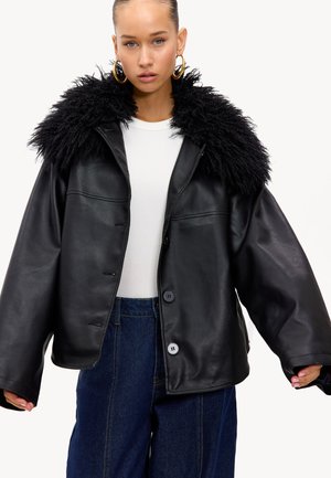 Jeune femme portant une grande veste en cuir noire avec un large col en fourrure, un haut blanc, des boucles d'oreilles dorées et un jean bleu.