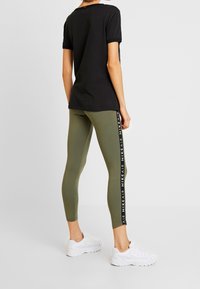 Musta t-paita yhdistettynä oliivinvihreisiin legginsseihin, joissa on valkoinen sivukaistale ja "NIKE AIR" -teksti, täydentäen asua valkoisilla lenkkareilla.