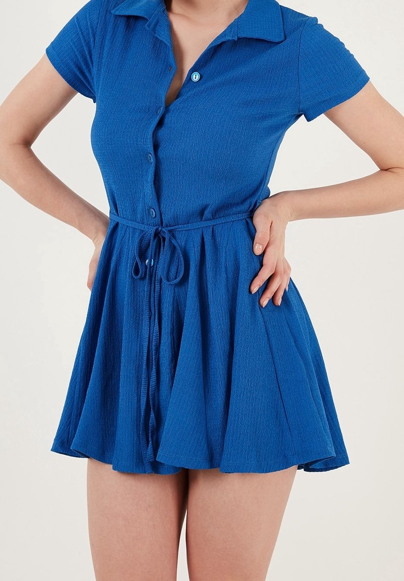 Kobieta w krótkiej, niebieskiej sukience na guziki z kołnierzykiem i paskiem w talii, z dłońmi opartymi na biodrach, na jednolitym tle.