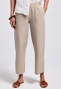 Person som bär beige och vita vertikala randiga byxor, en vit topp, bruna slidesandaler och ett mörkbrunt armband på vänster handled.