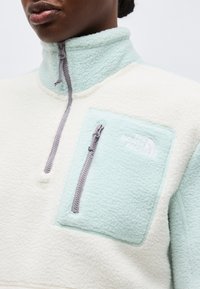 Zbliżenie osoby noszącej biało-niebieską polarkę z kieszenią na piersi na zamek i wysokim kołnierzem zapinanym do połowy.