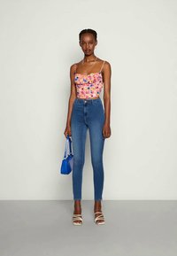 Top cropped rosa com padrão floral e detalhes em renda, combinado com jeans skinny azuis de cintura alta e sandálias brancas de salto. Acessório de bolsa azul.