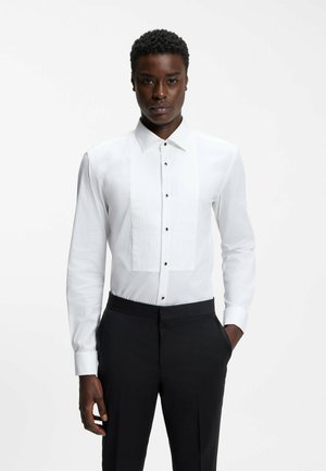 Man wearing a fitted white dress shirt with pleated front panels and black dress pants, standing against a plain background.