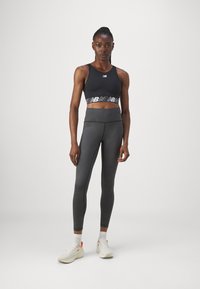 Fekete sportmelltartó logós pánttal, sötétszürke, magas derekú leggingsszel párosítva. A modellen fehér sportcipők vannak piros és sárga részletekkel.