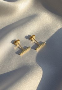 Boucles d'oreilles en or avec un design de barre minimaliste, des surfaces lisses et des fermoirs papillon, reposant sur un tissu doux et scintillant.
