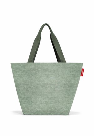 Grüne Stofftragetasche mit konischer Form, zwei flachen Griffen und einem kleinen roten Logosticker. Strukturierter Stoff mit einer weichen Oberfläche.