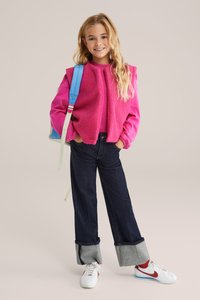 Ružová fleecová bunda s rebrovanými manžetami, v kombinácii s tmavými džínsami s obrúbeným lemom. Biele tenisky s červenými akcentmi dopĺňajú outfit.