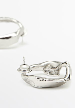 Silberohrringe mit abstrakter, organischer Form. Einer davon weist einen kleinen glockenförmigen Akzent auf, der eine glatte, reflektierende Oberfläche betont.