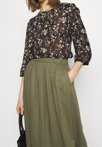 Transparent svart blus med färgglad blommönster, kombinerad med en högmidjad olivgrön kjol med veck och fickor. Svart handväska hålls i handen.