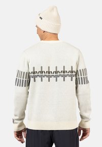 Hellgrauer gestrickter Pullover mit schwarzen geometrischen Mustern auf dem Rücken und den Ärmeln, ausgestattet mit einem gerippten Saum und Bündchen. Kombiniert mit einer beigen Mütze.