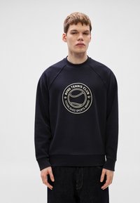 Tumši zils sporta džemperis ar apaļu logo, kurā attēlots teksts "BOSS TENNIS CLUB" un tenisa bumbiņas grafika; izgatavots no mīksta auduma ar ribotiem aproču galiem.