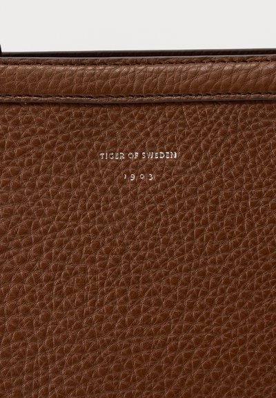Καφέ δερμάτινη επιφάνεια με υφή και ανάγλυφο λευκό κείμενο "Tiger of Sweden 1903" κάτω από τη ραμμένη άκρη.