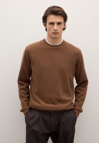Pull en maille marron avec un col rond et des manches longues, présentant une texture douce et une coupe décontractée, associé à un pantalon foncé.