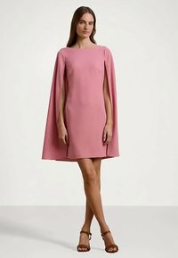 Femeie îmbrăcată într-o rochie cu pelerină roz, cu mâneci deschise, purtând pantofi cu toc deschis, de culoare maronie, pe un fundal simplu de culoare deschisă.