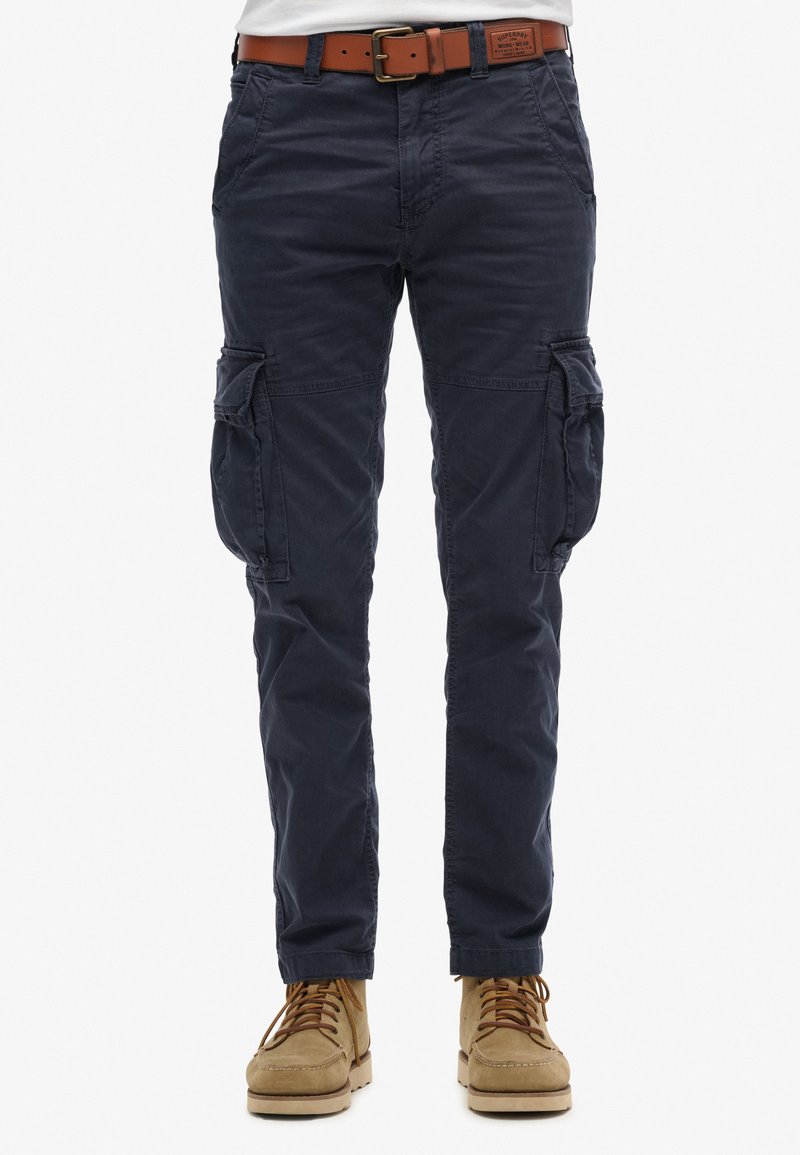 Pantalones cargo en azul marino con un ajuste relajado, presentan bolsillos laterales, detalles cosidos y un cinturón de cuero en tono marrón. Tejido texturizado.