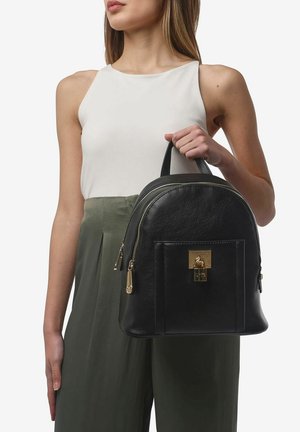 Zaino in pelle nera con forma arrotondata, hardware dorato, tasca frontale con chiusura e due cerniere laterali, indossato da un modello.