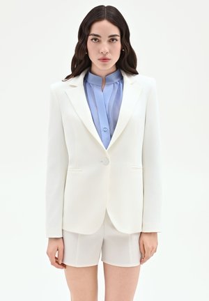 Donna con lunghi capelli scuri indossa blazer bianco e pantaloncini abbinati sopra una camicia azzurro chiaro con colletto su sfondo bianco.