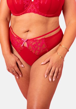 Frau trägt rotes besticktes Dessous-Set mit dekorativen Trägern, rosa Schleife an der Unterhose und goldenen Armbändern am rechten Handgelenk.