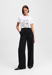 Witte t-shirt met zwarte grafische print, gecombineerd met zwarte high-waisted wijde broek. Model staat met handen in de zakken en draagt zwarte schoenen.