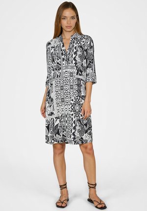 Woman wearing a black and white patterned knee-length dress with rolled-up sleeves and black strappy sandals, standing against a white background.