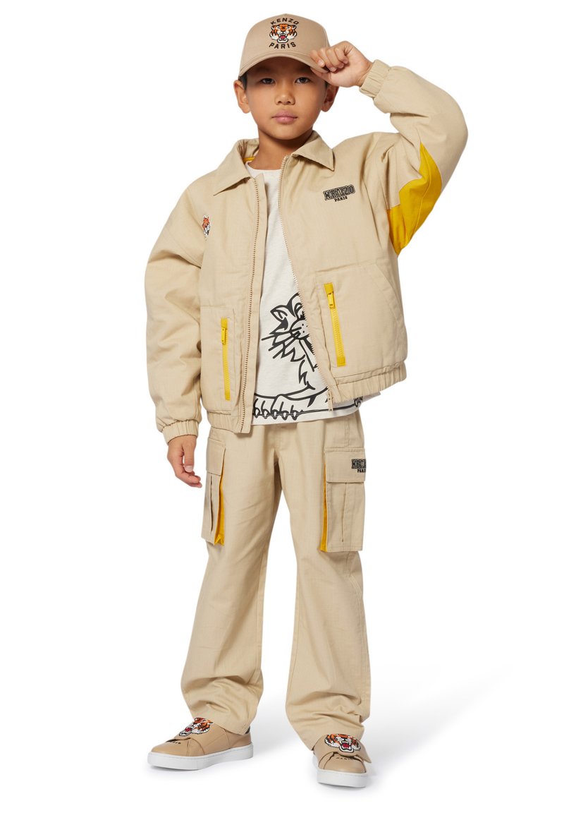 Veste beige avec des accents jaunes, t-shirt blanc avec un contour de lion, pantalon cargo et baskets beiges avec un design de tigre. La casquette assortie à la veste.