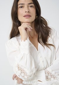 White blouse with long, puffy sleeves featuring lace accents and a button-down front. Soft texture and intricate lace detailing around the neckline.