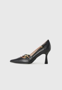 Chaussure à talon haut en cuir noir avec un bout pointu, présentant des accents en métal doré et un design découpé sur les côtés.