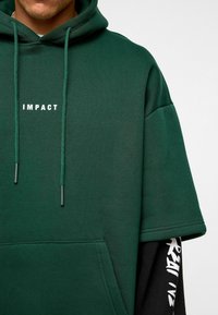 Sweat à capuche vert avec le mot "IMPACT" en blanc. Il possède une poche kangourou, des cordons de serrage et des manches courtes, superposé sur un haut à manches longues noir.