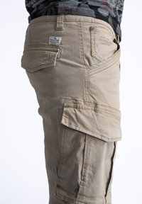 Pantalones cargo beige con varios bolsillos laterales, corte recto, costuras visibles y una pequeña etiqueta de la marca en el bolsillo trasero.