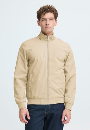 Młody mężczyzna o kręconych włosach, ubrany w beżową kurtkę na zamek oraz ciemnoniebieskie jeansy, stojący na tle jednolitego jasnego koloru.