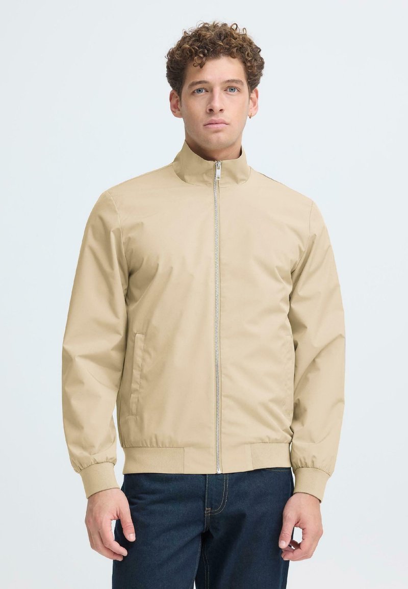 Ung mand med krøllet hår iført en beige lynlåstrøje og mørkeblå jeans, stående mod en ensfarvet lys baggrund.