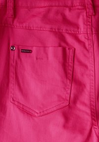 Tissu rose vif avec une texture légèrement brillante, présentant une petite poche arrière et une étiquette en métal. Des coutures visibles soulignent les bords.