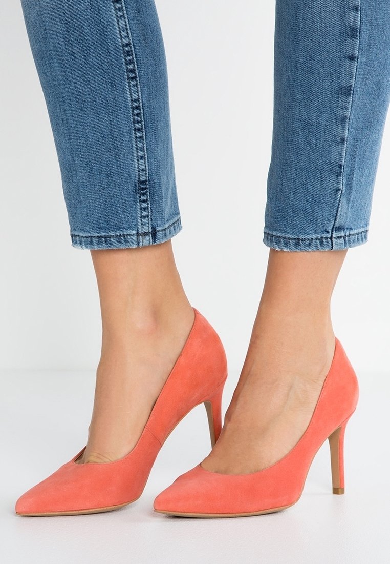 Pieds chaussés de talons hauts pointus en daim corail associés à un jean en denim bleu coupé au-dessus de la cheville, sur un fond uni.